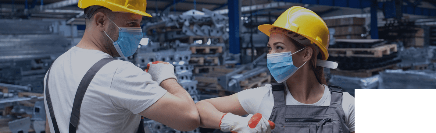 Two factory workers wearing hard hats, face masks, and gloves greet each other with an elbow bump in an industrial warehouse setting showcasing career