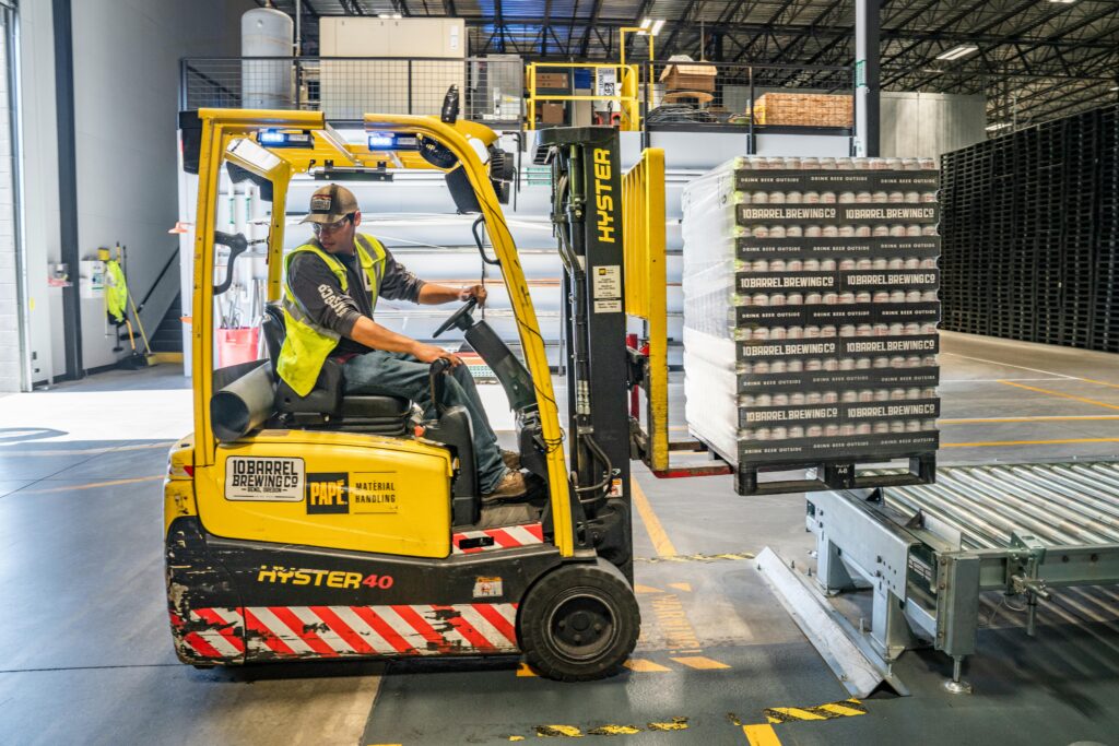 Why Do Manufacturers Need An MES? A supply chain worker moving boxes in the warehouse on a yellow fork lifter truck. Understanding MES implementation for manufacturers.